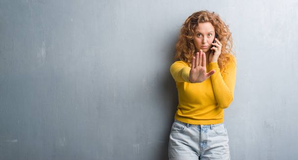 Young redhead woman over grey grunge wall talking on the phone with open hand doing stop sign with serious and confident expression, defense gesture - Φωτογραφία, εικόνα