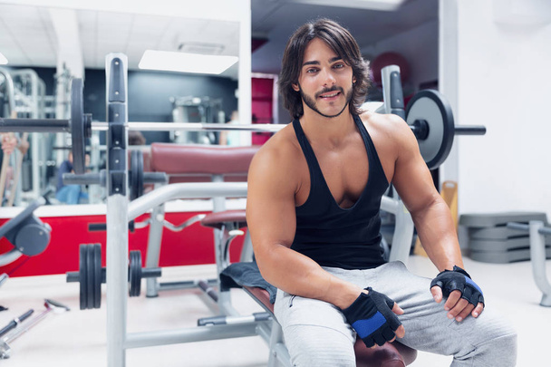 Porträt eines jungen erwachsenen Bodybuilders, der auf einer Bank in einem Fitnessstudio sitzt und der Kamera ein fröhliches freundliches Lächeln schenkt; Konzept für Gesundheit und Fitness - Foto, Bild