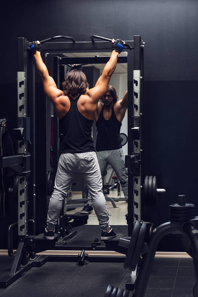 Αθλητική μυώδης άνδρας εργάζεται έξω κάνει pull-ups σε ένα γυμναστήριο που παρατηρείται από πίσω, να ενισχύσει τους μυς του - Φωτογραφία, εικόνα