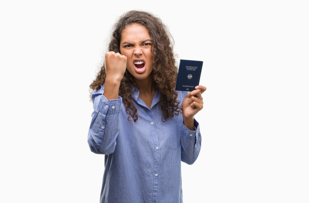 Young hispanic woman holding passport of Germany annoyed and frustrated shouting with anger, crazy and yelling with raised hand, anger concept - Photo, Image
