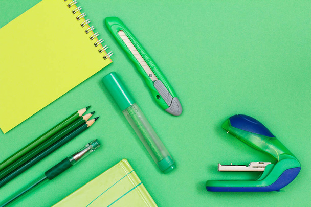 Notebook, color pencils, pen, book, felt-tip pen, paper knife and stapler on green background. Top view. Back to school concept. School supplies - 写真・画像