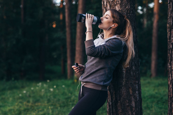 Nainen lenkkeilijä toipumassa jälkeen intensiivinen harjoitus seisoo lähellä puun juomaveden, yllään kuulokkeet puistossa
 - Valokuva, kuva