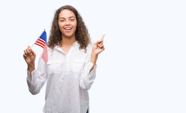 Young hispanic woman holding flag of United Estates of America very happy pointing with hand and finger to the side - Φωτογραφία, εικόνα