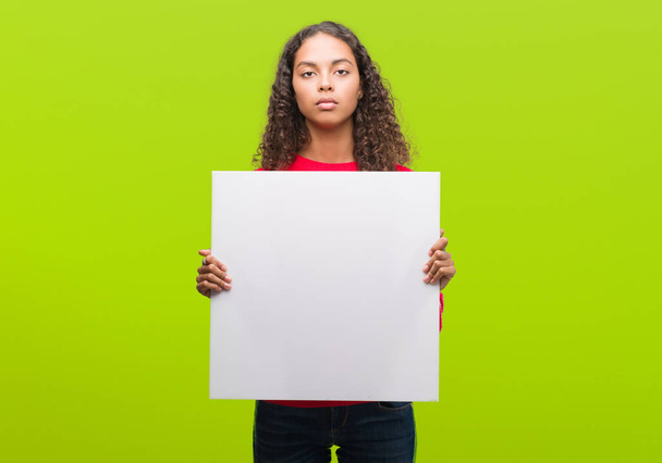 Young hispanic woman holding blank banner with a confident expression on smart face thinking serious - Photo, Image