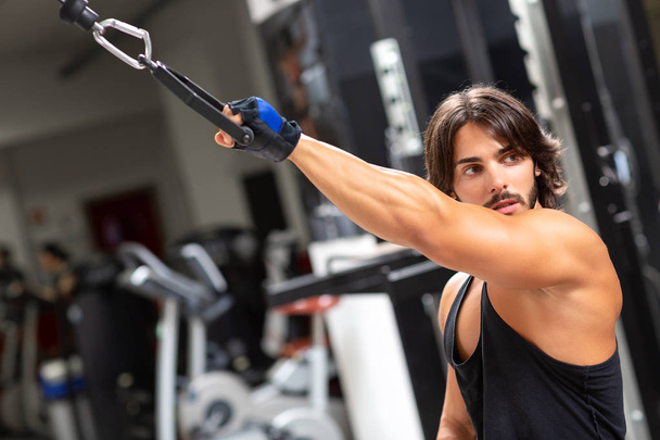 Single bearded adult male bodybuilder pulling black ring attached to machine off camera with one hand - Foto, Bild