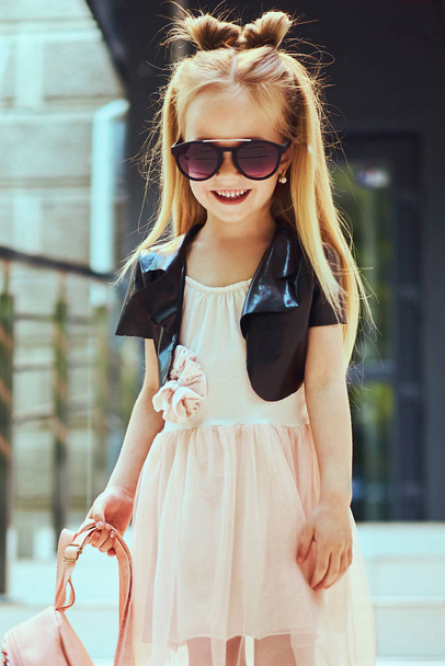 Little girl child wearing a sunglasses and backpack standing in over city background - Фото, изображение