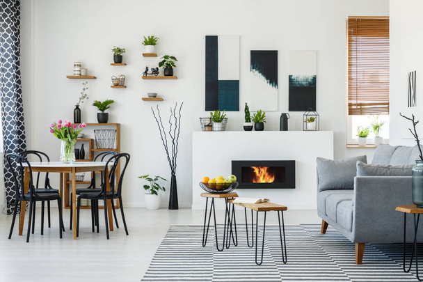 Chairs at table with flowers in white apartment interior with grey sofa next to fireplace. Real photo - Valokuva, kuva