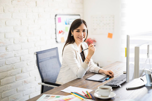 Портрет улыбающейся женщины-дизайнера, сидящей за столом с графическим планшетом и компьютером в офисе
 - Фото, изображение