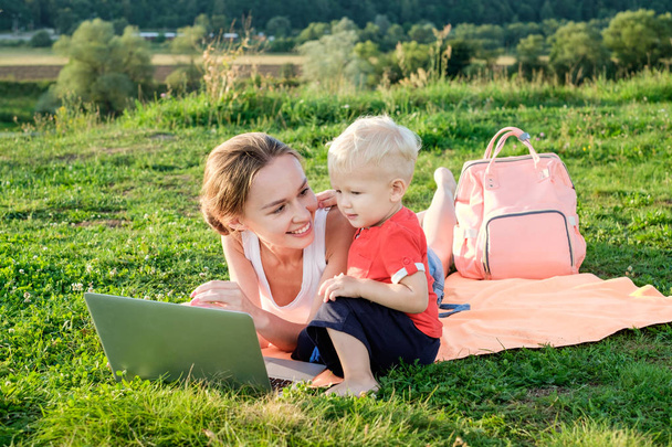 Mather and baby lie on green grass on a plaid, communicate, smile, in front of a laptop - Valokuva, kuva