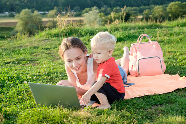 Mather and baby lie on green grass on a plaid, communicate, smile, in front of a laptop - Fotoğraf, Görsel