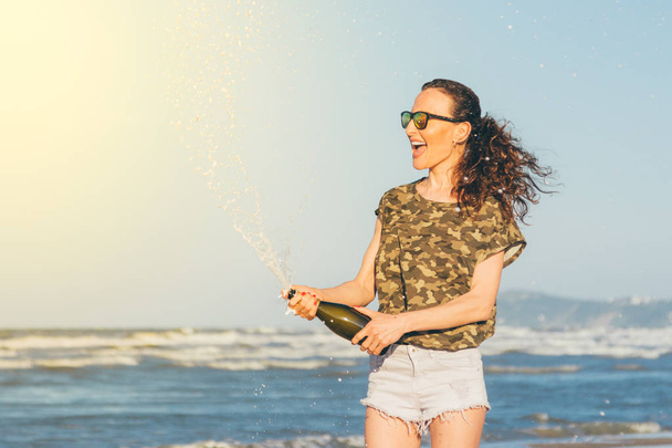 Pretty young woman splashing champagne on the beach against the blue sky and a stormy sea - Fotografie, Obrázek