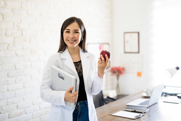 Retrato de jovem nutricionista feminina sorridente segurando balança de peso e maçã na sala de consulta
 - Foto, Imagem