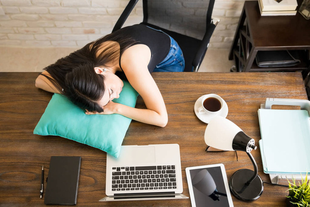 Blick aus der Vogelperspektive auf junge Frau, die mit Kissen an ihrem Schreibtisch schläft - Foto, Bild