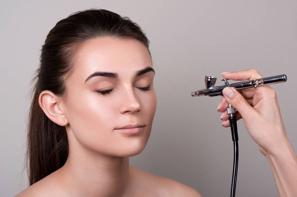 Makeup artist using airbrush. Fashion brunette model with hair tail over gray background. Professional makeup with aerograph. Girl doing makeup in beauty salon. Female face with natural clean skin - Fotó, kép