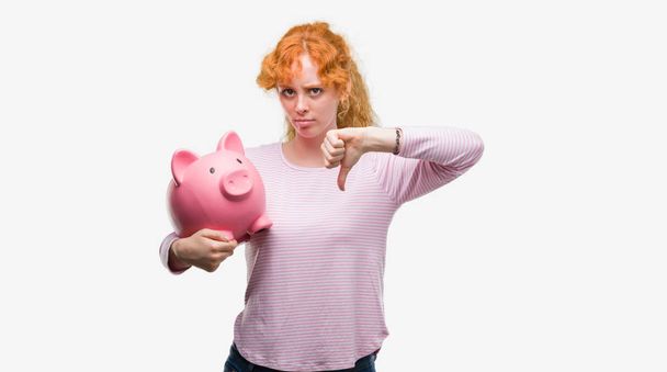 Fiatal vöröshajú nő gazdaság malacka bank dühös arccal, negatív előjellel mutatja nem tetszik a hüvelykujját lefelé, elutasítás koncepció - Fotó, kép