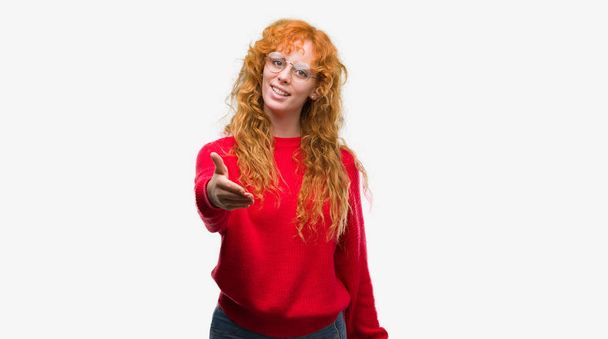 Młoda Ruda kobieta sobie czerwony sweter uśmiechający się oferujący przyjazny uścisk dłoni jako pozdrowienie i przyjazna. Udanego biznesu. - Zdjęcie, obraz