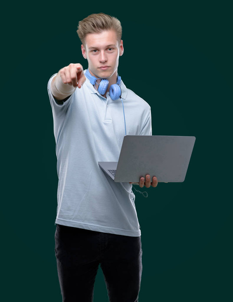 Knappe blonde jongeman met behulp van een laptop met de vinger te wijzen naar de camera en u, hand teken, positieve en zelfverzekerd gebaar van de voorkant - Foto, afbeelding