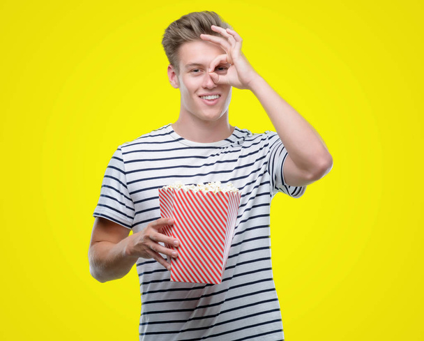 Mladý pohledný blondýn jíst popcorn s šťastný obličej s úsměvem dělá ok znamení rukou na oko dívá skrze prsty - Fotografie, Obrázek