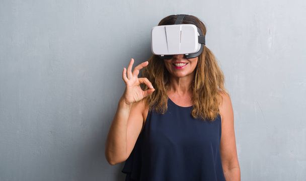 hispanische Frau mittleren Alters, die mit Virtual-Reality-Brille über der grauen Grunge-Wand steht und mit den Fingern Okay-Zeichen macht, hervorragendes Symbol - Foto, Bild