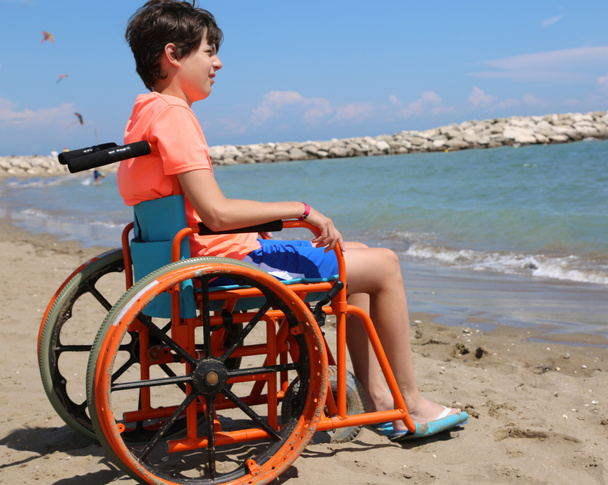 νεαρό αγόρι σχετικά με το ειδικό αναπηρικό αμαξίδιο με μεγάλους τροχούς να κινείται ομαλά πάνω στην άμμο της παραλίας δίπλα στη θάλασσα το καλοκαίρι - Φωτογραφία, εικόνα