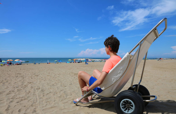 αγόρι σε ένα ειδικό αναπηρικό αμαξίδιο να κινηθεί ομαλά στην αμμώδη παραλία, δίπλα στη θάλασσα το καλοκαίρι - Φωτογραφία, εικόνα