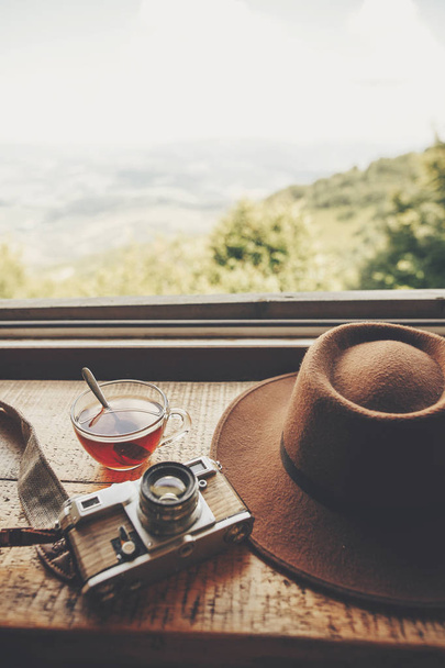 zomer reizen en wanderlust. stijlvolle hipster hoed, fototoestel en thee in glas met lepel op houten tafel in venster licht met uitzicht op de bergen en de lucht. heerlijke warme drank op de top - Foto, afbeelding