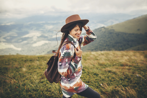 travel and wanderlust concept. stylish traveler hipster girl holding hat, with backpack and windy hair, walking in mountains in clouds. summer vacation. space for text. amazing atmospheric moment - Photo, Image