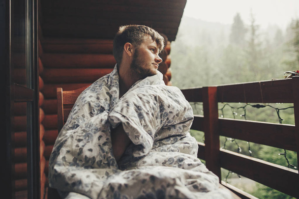 stijlvolle bebaarde man ontspannen op houten veranda onder bos in Regenachtig Bergen. de kerel van de Hipster rusten in deken, zittend in huisje, kijken naar regen. rustige vreedzame moment - Foto, afbeelding