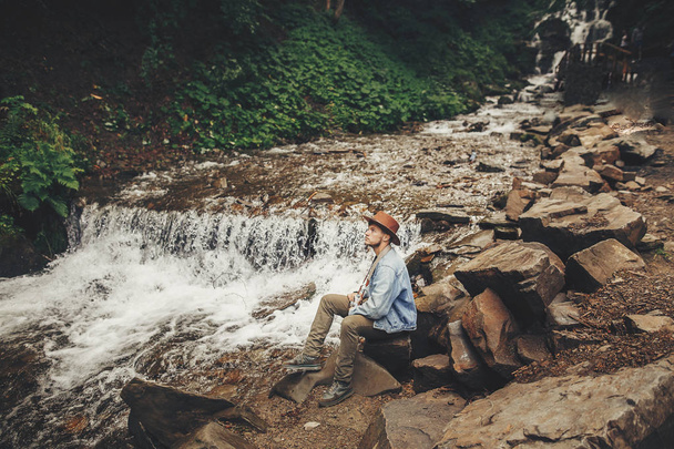 stylischer Hipster mit Hut und Fotokamera, der am Fluss mit Wasserfall im Wald in den Bergen sitzt. Reisender, der Wälder erkundet. Raum für Text. Reise- und Fernweh-Konzept. - Foto, Bild
