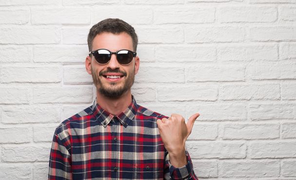 Молодий дорослий чоловік у сонцезахисних окулярах стоїть над білою цегляною стіною, вказуючи і показуючи з великим пальцем в сторону з щасливим обличчям посміхаючись
 - Фото, зображення