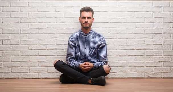 Νεαρός ενήλικος άνδρας κάθεται πάνω από το λευκό τούβλο τοίχο σκεπτικιστής και νευρικό, frowning αναστατωμένος εξαιτίας προβλήματος. Αρνητικό πρόσωπο. - Φωτογραφία, εικόνα