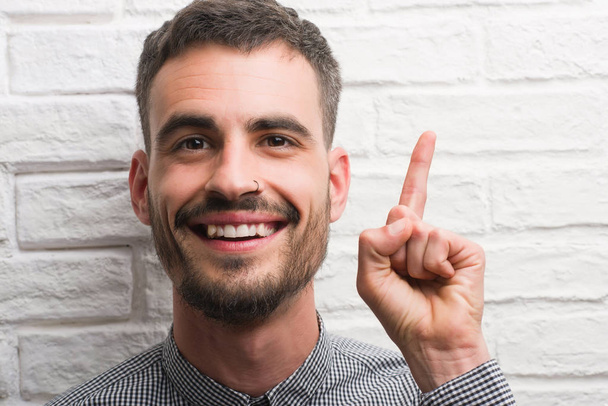 Hombre adulto joven de pie sobre la pared de ladrillo blanco sorprendido con una idea o pregunta que apunta el dedo con la cara feliz, número uno
 - Foto, imagen