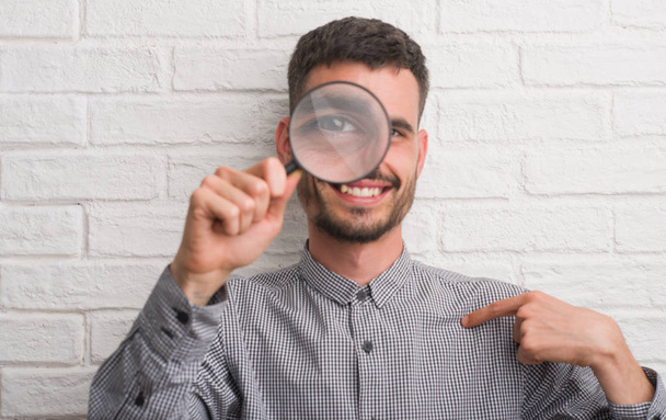 Young adult man over brick wall using magnifying glass with surprise face pointing finger to himself - Photo, Image