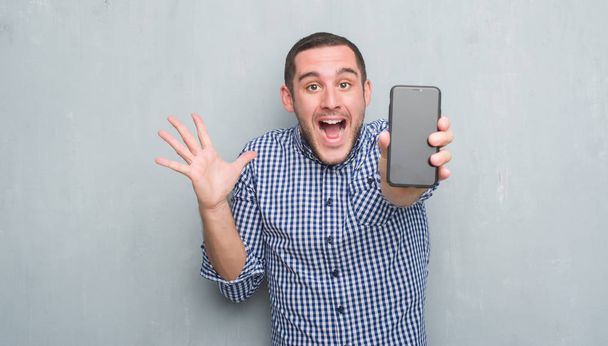 Young caucasian man over grey grunge wall showing blank screen of smartphone very happy and excited, winner expression celebrating victory screaming with big smile and raised hands - Foto, Imagen