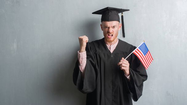 Jeune homme rousse sur le mur gris grunge portant uniforme diplômé tenant drapeau américain ennuyé et frustré criant de colère, fou et criant avec la main levée, concept de colère
 - Photo, image