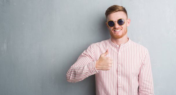 Νέοι κοκκινομάλλα άνδρα πάνω σε γκρι grunge τοίχου φορώντας retro γυαλιά ηλίου ευτυχισμένος με μεγάλο χαμόγελο κάνει εντάξει σήμα, τον αντίχειρα επάνω με τα δάχτυλα, καλό σημάδι - Φωτογραφία, εικόνα
