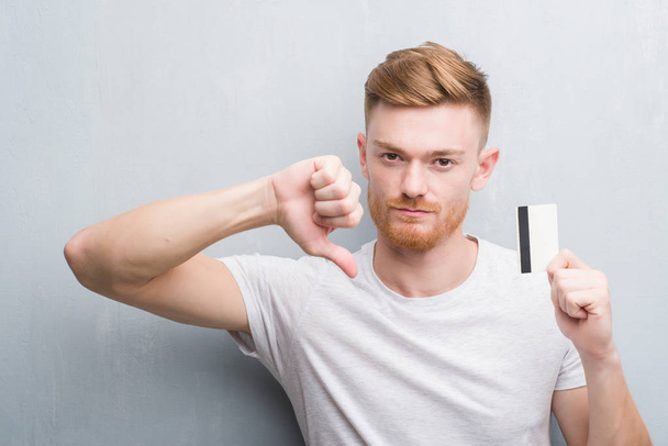 Mladá zrzka muž přes zeď šedá grunge drží kreditní kartu s naštvaný obličej, záporné znaménko ukazující nechuť s palci dolů, koncept odmítnutí - Fotografie, Obrázek