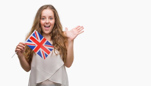 Mladá blond žena hospodářství vlajka Spojeného království velmi šťastný a nadšený, vítěz výrazu slaví vítězství s křikem velký úsměv a pozvedl ruce - Fotografie, Obrázek