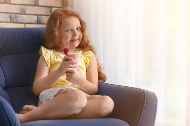 Little girl with glass of delicious milk shake in armchair indoors - 写真・画像