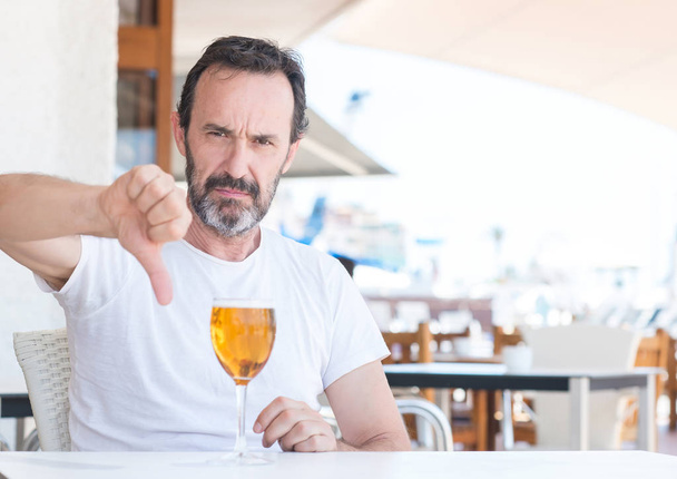 Szép magas rangú ember iszik sört, haragos arc, negatív előjellel mutatja ellenszenv a hüvelykujját lefelé, elutasítás koncepció étteremben - Fotó, kép