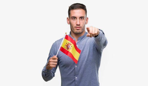 Jovem bonito segurando uma bandeira da Espanha apontando com o dedo para a câmera e para você, sinal de mão, gesto positivo e confiante da frente
 - Foto, Imagem