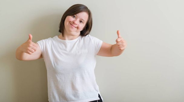 Frau mit Down-Syndrom, die über einem Erfolgsschild steht und eine positive Geste mit der Hand macht, die Daumen hoch lächelnd und glücklich. Blick in die Kamera mit heiterem Gesichtsausdruck, Siegergeste. - Foto, Bild