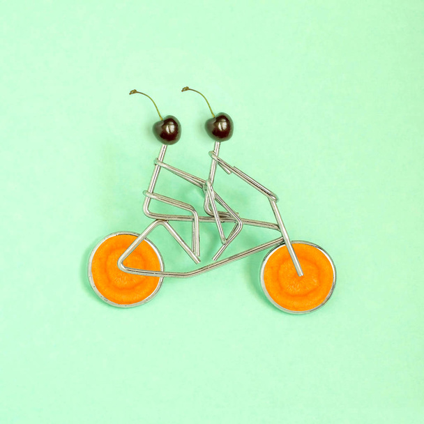 Metallfiguren von Sportlern mit Kirschkopf auf Fahrrad mit Karottenrädern auf grünem Pastellgrund. Minimaler Stil. Konzept der gesunden Lebensweise, Diät- und Sportkost - Foto, Bild