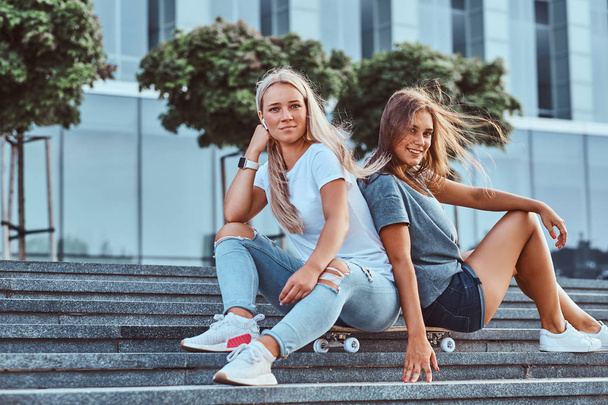 Портрет двох молодих дівчат-гіпстерів, що сидять разом на скейтборді на сходинках на тлі хмарочоса
. - Фото, зображення