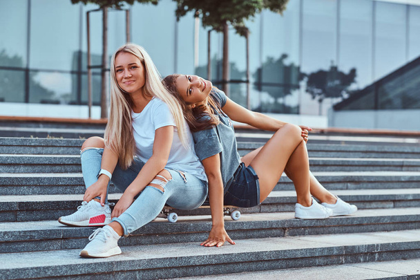 高層ビルの背景にステップでスケート ボードに一緒に座っている二人の流行に敏感な若い女の子の肖像画. - 写真・画像