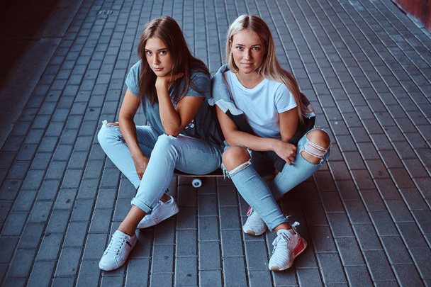 Δύο όμορφες hipster κορίτσια κάθονται για skateboard στο πεζοδρόμιο κάτω από γέφυρα. - Φωτογραφία, εικόνα