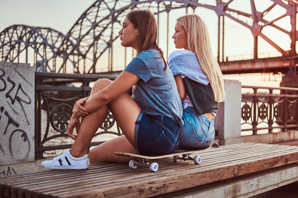 A két fiatal hipster lányok ült egy gördeszka és nézett el a háttérben a régi híd sunset oldalnézetből. - Fotó, kép