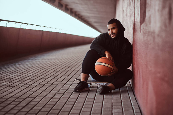 Lächelnder dunkelhäutiger Typ in schwarzem Kapuzenpulli und Sporthose hält einen Basketball in der Hand, während er auf einem Skateboard sitzt und sich an eine Wand lehnt. - Foto, Bild