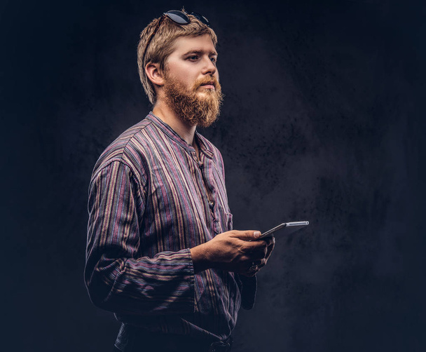 Redhead bearded hipster guy dressed in an old-fashioned shirt holds a tablet computer. Isolated on a dark background. - Photo, Image