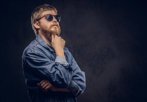 Pensif hipster gars portant des lunettes de soleil vêtu d'une veste en jeans posant avec la main sur le menton sur un fond sombre
. - Photo, image